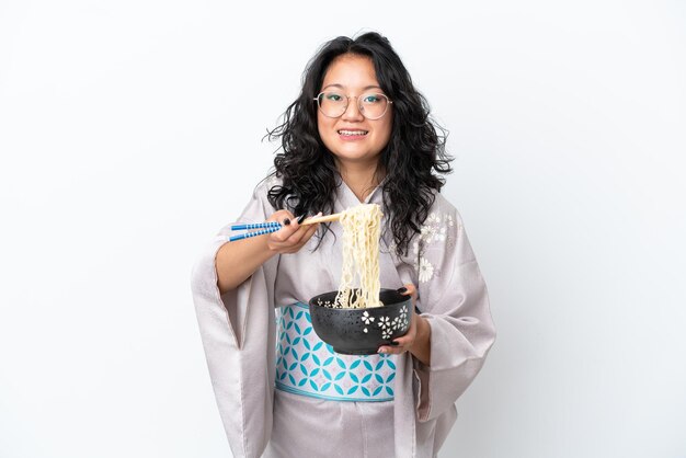 Joven asiática vistiendo kimono aislado sobre fondo blanco sosteniendo un plato de fideos con palillos y ofreciéndolo