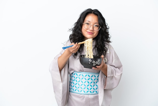 Joven asiática vistiendo kimono aislado sobre fondo blanco sosteniendo un plato de fideos con palillos