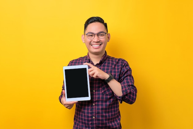 Un joven asiático alegre con anteojos que muestra una tableta digital con una pantalla vacía para una maqueta aislada sobre un fondo amarillo