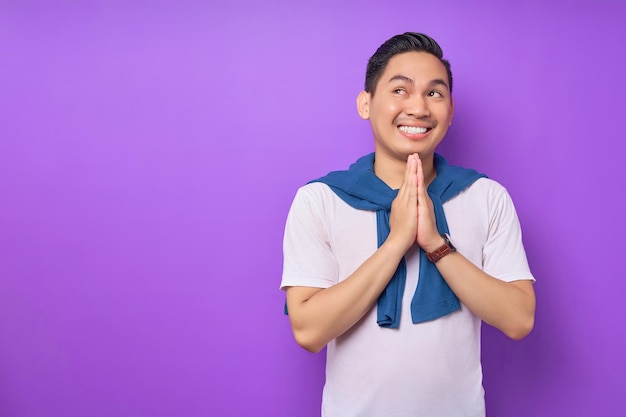 Joven asiático alegre con camiseta blanca colocando las manos en gesto de oración y mirando a un lado aislado sobre el concepto de estilo de vida de la gente de fondo púrpura