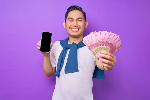 Un joven asiático alegre con una camiseta blanca que muestra un teléfono móvil con pantalla en blanco y tiene billetes de dinero aislados en un fondo morado