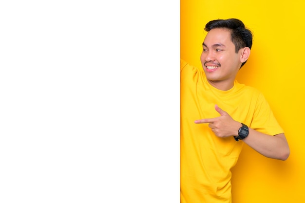 Un joven asiático alegre con una camiseta informal apuntando a un tablero de anuncios blanco aislado en un fondo amarillo Concepto de cartelera de promoción