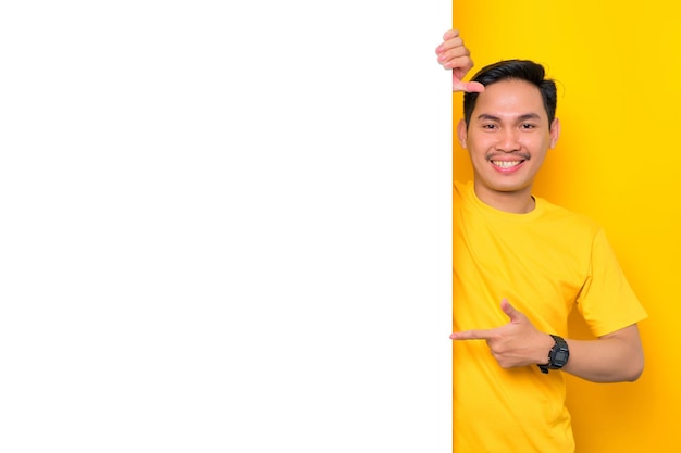 Un joven asiático alegre con una camiseta informal apuntando a un tablero de anuncios blanco aislado en un fondo amarillo Concepto de cartelera de promoción