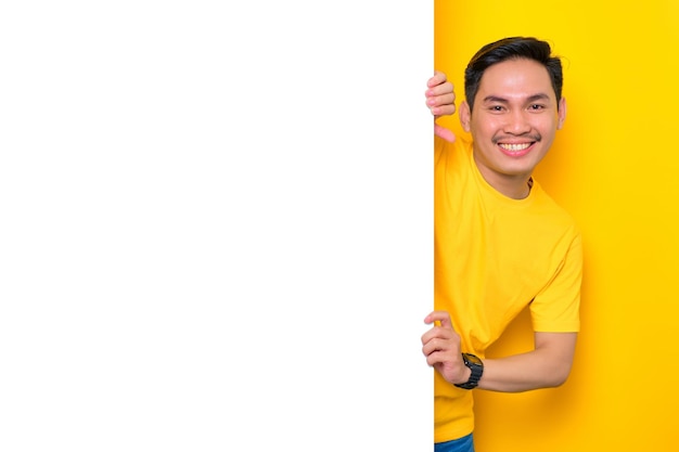 Un joven asiático alegre con una camiseta informal asomándose al lado de un tablero de anuncios blanco aislado en un fondo amarillo Concepto de cartelera de promoción