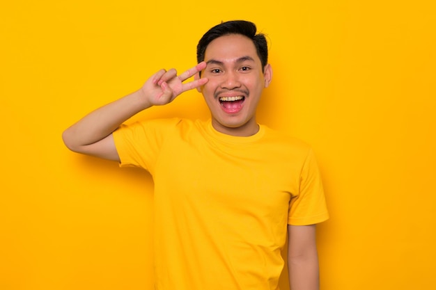 Un joven asiático alegre con una camiseta informal que muestra el signo de la paz con los dedos sobre la cara aislado en un fondo amarillo Concepto de estilo de vida de la gente