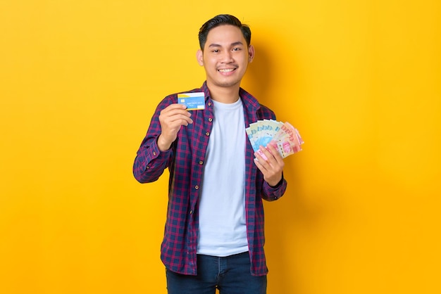 Un joven asiático alegre con pantalones a cuadros sosteniendo billetes de dinero y mostrando una tarjeta de crédito aislada de fondo amarillo