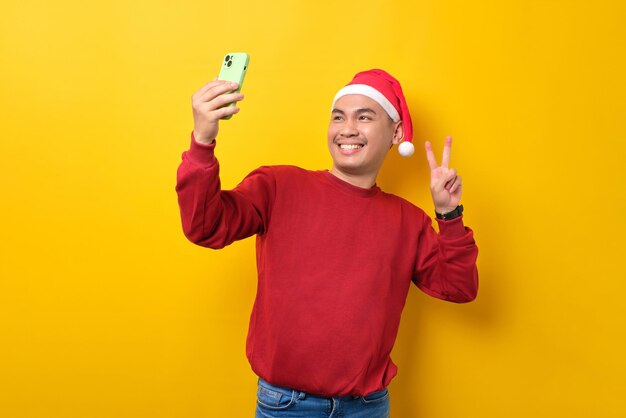 Un joven asiático alegre con sombrero de Papá Noel tomándose un selfie en un smartphone mostrando el signo de la victoria sobre la celebración de fondo del estudio amarillo, las vacaciones de Navidad y el concepto de Año Nuevo