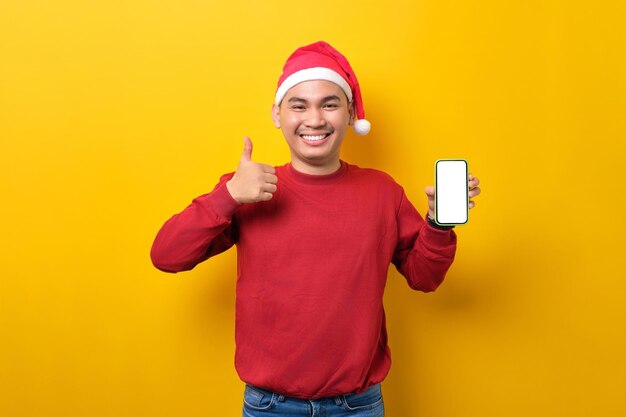 Un joven asiático alegre con sombrero de Santa sosteniendo un teléfono móvil con una pantalla en blanco que muestra un gesto con el pulgar hacia arriba sobre el fondo amarillo del estudio, la celebración de las vacaciones de Navidad y el concepto de Año Nuevo