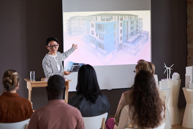 Joven asiático en anteojos apuntando a la pizarra con edificio moderno y explicando el plan para gente de negocios en el seminario