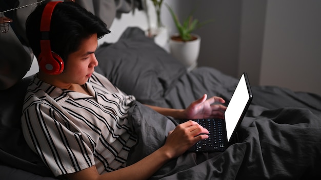 Joven asiático con auriculares y navegar por internet con tableta en su cama.