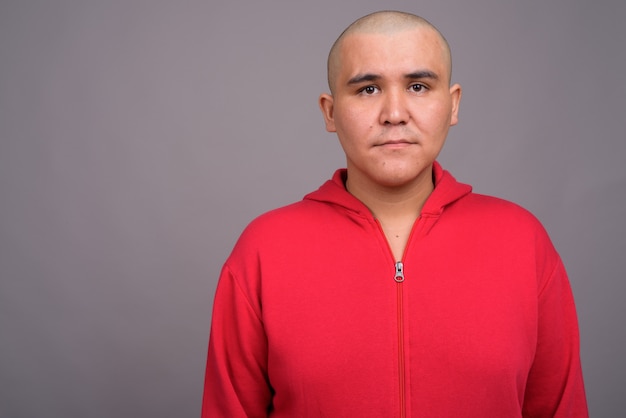 joven asiático calvo vistiendo chaqueta roja contra la pared gris