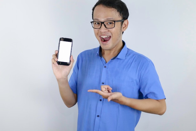 Un joven asiático con camisa azul está de pie y sonriendo señalando un espacio en blanco en la pantalla del teléfono inteligente con fondo blanco