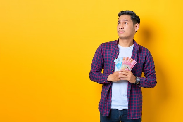 Joven asiático con camisa a cuadros sosteniendo billetes de dinero y mirando hacia otro lado en el espacio de copia aislado en el fondo amarillo