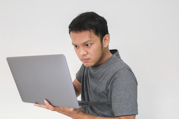 Joven asiático con camiseta gris que se siente serio y se concentra en sostener la computadora portátil en la mano