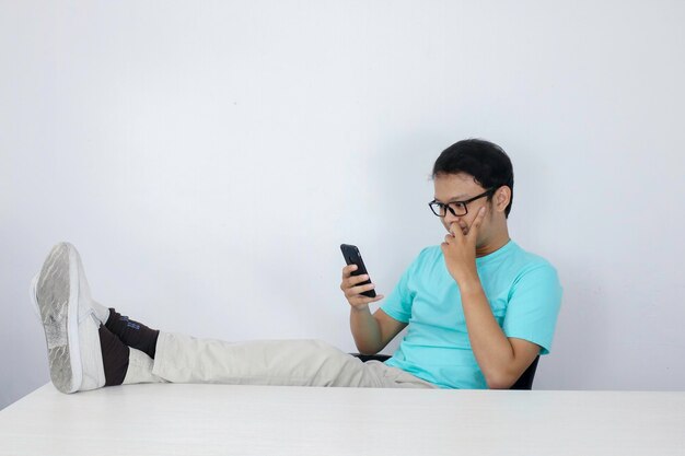 Joven asiático con cara feliz con lo que ve en un teléfono móvil con la pierna sobre la mesa Hombre indonesio con camisa azul