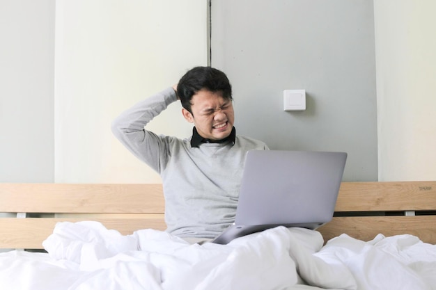 El joven asiático con una computadora portátil en la cama está confundido y estresado Concepto de trabajo desde casa