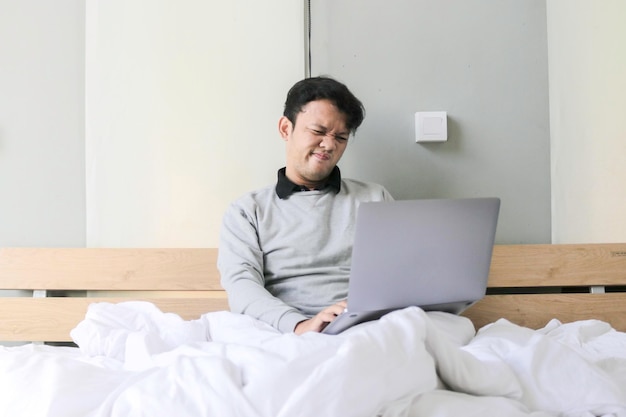El joven asiático con una computadora portátil en la cama está confundido y estresado Concepto de trabajo desde casa