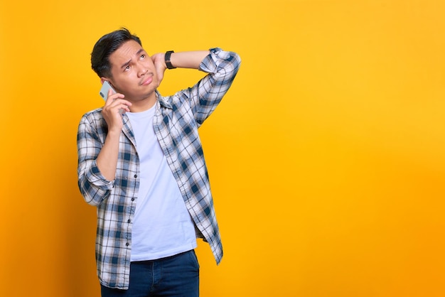 Joven asiático confundido con pantalones a cuadros hablando por teléfono móvil mirando a un lado el espacio de copia aislado en el fondo amarillo