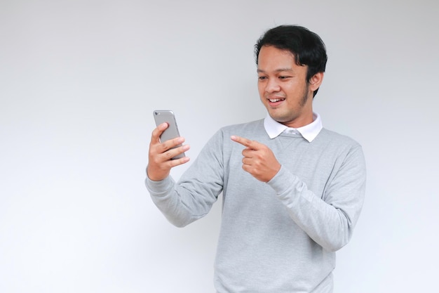 Joven asiático conmocionado y feliz con lo que ve en el teléfono inteligente en un fondo gris aislado