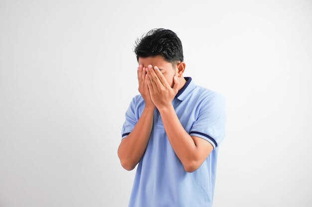 Un joven asiático deprimido se cubre la cara con las manos con una camiseta azul aislada de fondo blanco