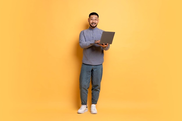 Joven asiático emocionado con una computadora portátil posando sobre un fondo de estudio amarillo