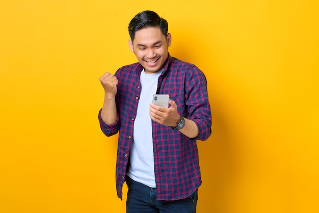 Un joven asiático emocionado con un pantalones a cuadros usando un smartphone y haciendo un gesto de ganador aislado en un fondo amarillo