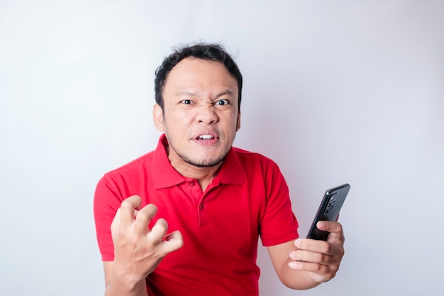 Un joven asiático enojado se ve descontento usando una camisa roja con expresiones faciales irritadas sosteniendo su teléfono