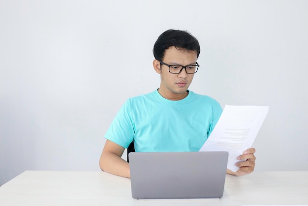 El joven asiático es serio y se enfoca cuando trabaja en una computadora portátil y documenta sobre la mesa. Hombre indonesio con camisa azul.