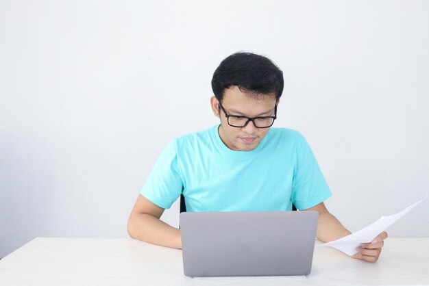 El joven asiático es serio y se enfoca cuando trabaja en una computadora portátil y documenta sobre la mesa. Hombre indonesio con camisa azul.