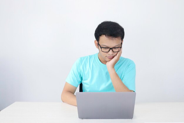 El joven asiático es serio y se enfoca cuando trabaja en una computadora portátil y documenta sobre la mesa. Hombre indonesio con camisa azul.