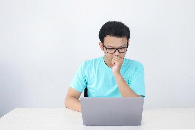 El joven asiático es serio y se enfoca cuando trabaja en una computadora portátil y documenta sobre la mesa. Hombre indonesio con camisa azul.