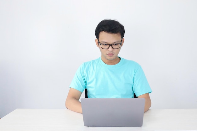 El joven asiático es serio y se enfoca cuando trabaja en una computadora portátil y documenta sobre la mesa. Hombre indonesio con camisa azul.