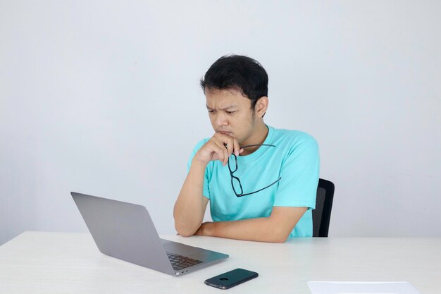 El joven asiático es serio y se enfoca cuando trabaja en una computadora portátil en la mesa. Hombre indonesio con camisa azul.