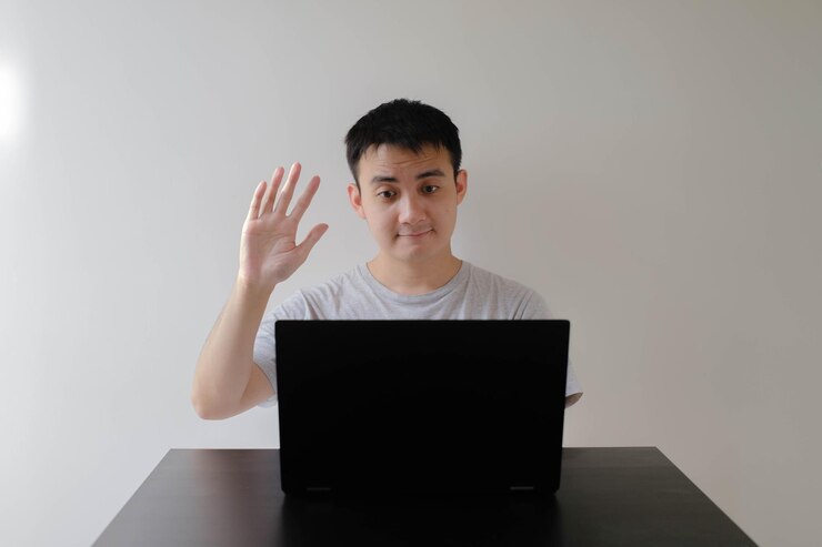 Un joven asiático está saludando con la mano a una reunión en línea en la computadora portátil