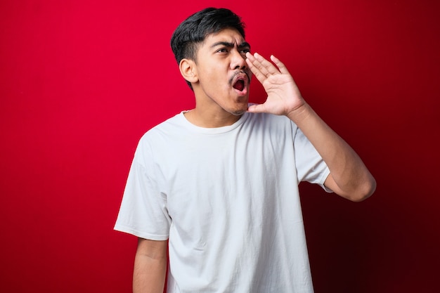 Joven asiático con estilo camiseta blanca gritando y gritando fuerte a un lado con la mano en la boca sobre fondo rojo. concepto de comunicación.