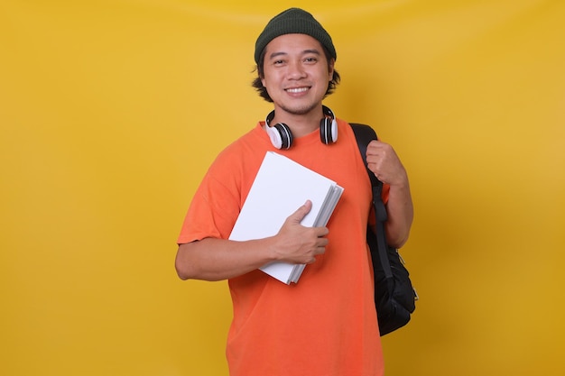 Joven asiático en estilo casual con mochila y auriculares trayendo libros aislados en backgr amarillo