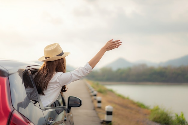 El joven asiático felicita el sombrero que lleva femenino dentro del coche negro compacto con levanta su mano a la ventanilla del coche con vistas a la montaña y al lago en un día soleado en Tailandia.