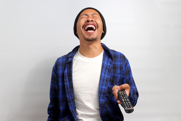 Un joven asiático feliz se ríe en voz alta mientras ve un programa de televisión hilarantemente divertido en casa