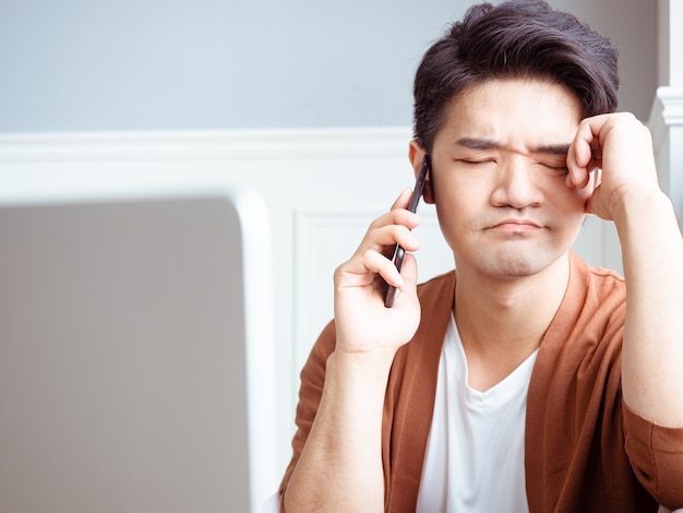 Joven asiático frotando sus ojos cansados con su teléfono inteligente.