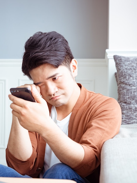 Joven asiático frotando sus ojos cansados con su teléfono inteligente.