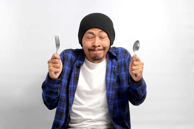 Un joven asiático hambriento imaginando una comida deliciosa con cuchara y tenedor