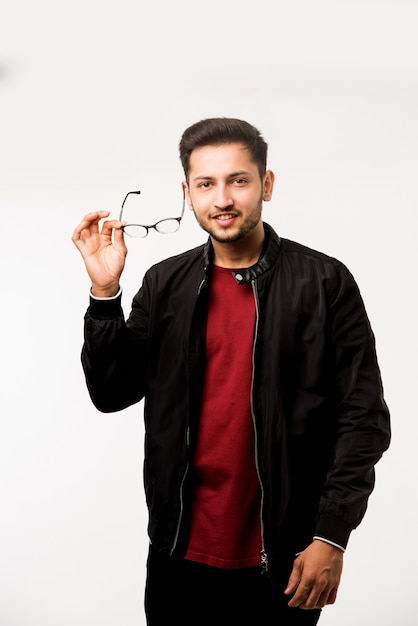 Joven asiático indio sosteniendo gafas o anteojos mientras está de pie sobre fondo blanco.