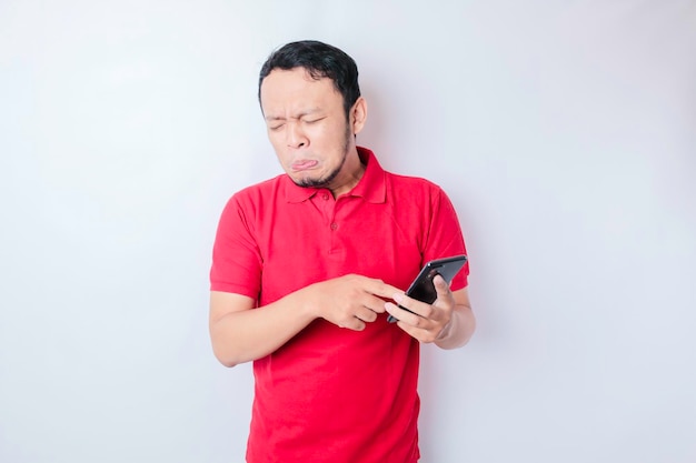 Un joven asiático insatisfecho parece descontento usando una camiseta roja con expresiones faciales irritadas sosteniendo su teléfono