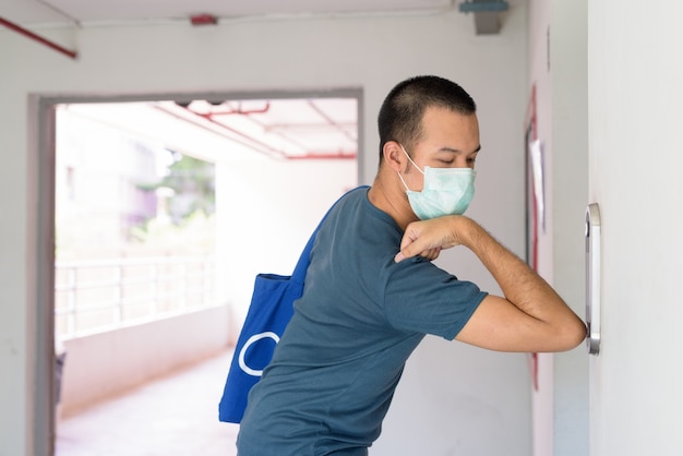 Joven asiático con máscara presionando el botón del elevador con el codo para prevenir la propagación del coronavirus