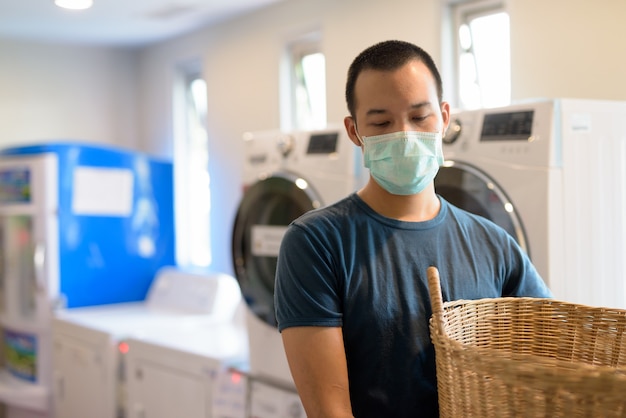 Joven asiático con máscara para protegerse del brote de coronavirus en la lavandería lavando ropa