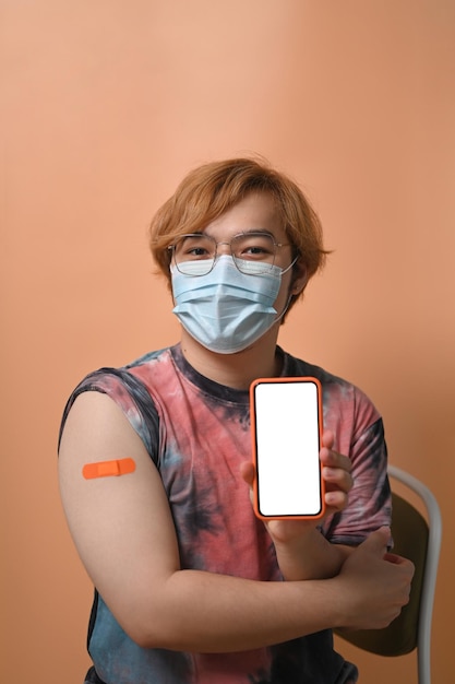 Joven asiático con mascarilla mostrando su brazo después de la vacunación y sosteniendo un teléfono inteligente