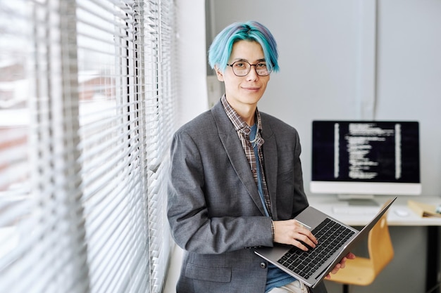 Joven asiático mirando a la cámara y sosteniendo una computadora portátil junto a la ventana