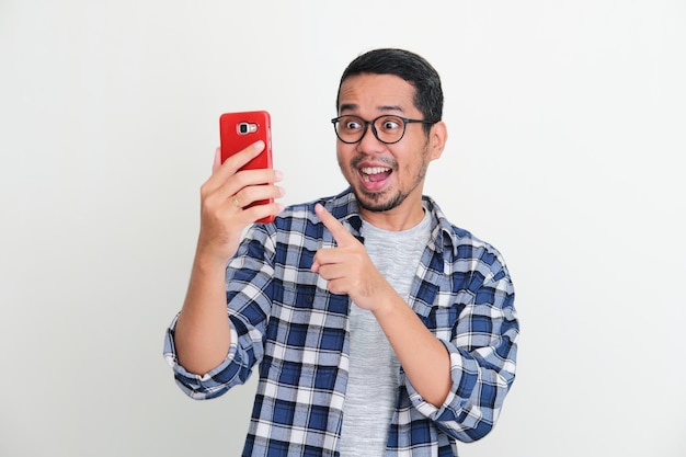 Joven asiático mirando y señalando su teléfono móvil con expresión sorprendida