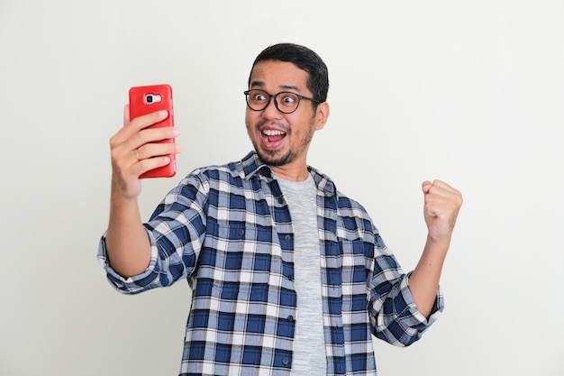 Joven asiático mostrando una expresión emocionada mientras mira su teléfono móvil