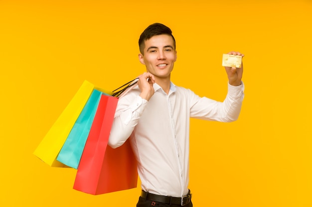 Joven asiático mostrando su bolsa de compras y tarjeta de crédito sobre fondo amarillo